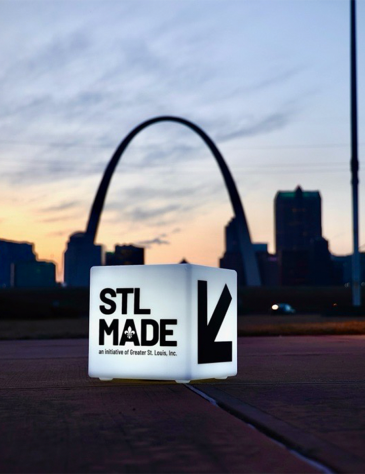  A luminous sign with "STL MADE" in front of the St. Louis Arch at dusk.