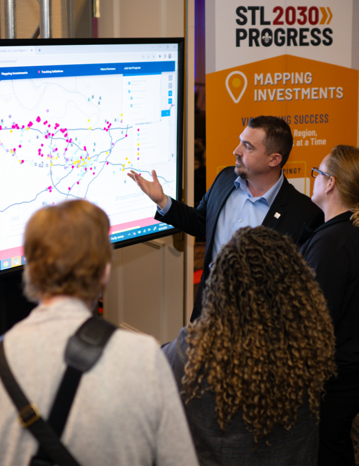 This image depicts a group of people gathered around a large screen displaying what appears to be a mapping application with various icons scattered across it, suggesting some sort of urban planning or investment analysis presentation. The participants are engaged in conversation, and one person seems to be explaining something related to the content on the screen. In the background, there’s a banner that reads “STL 2030 Progress” with a subtitle “Mapping Investments for Success."