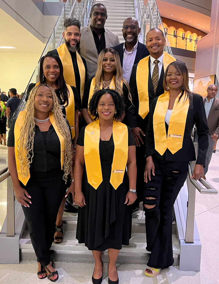A group of Fellows Experience graduates wearing all black with gold sashes.