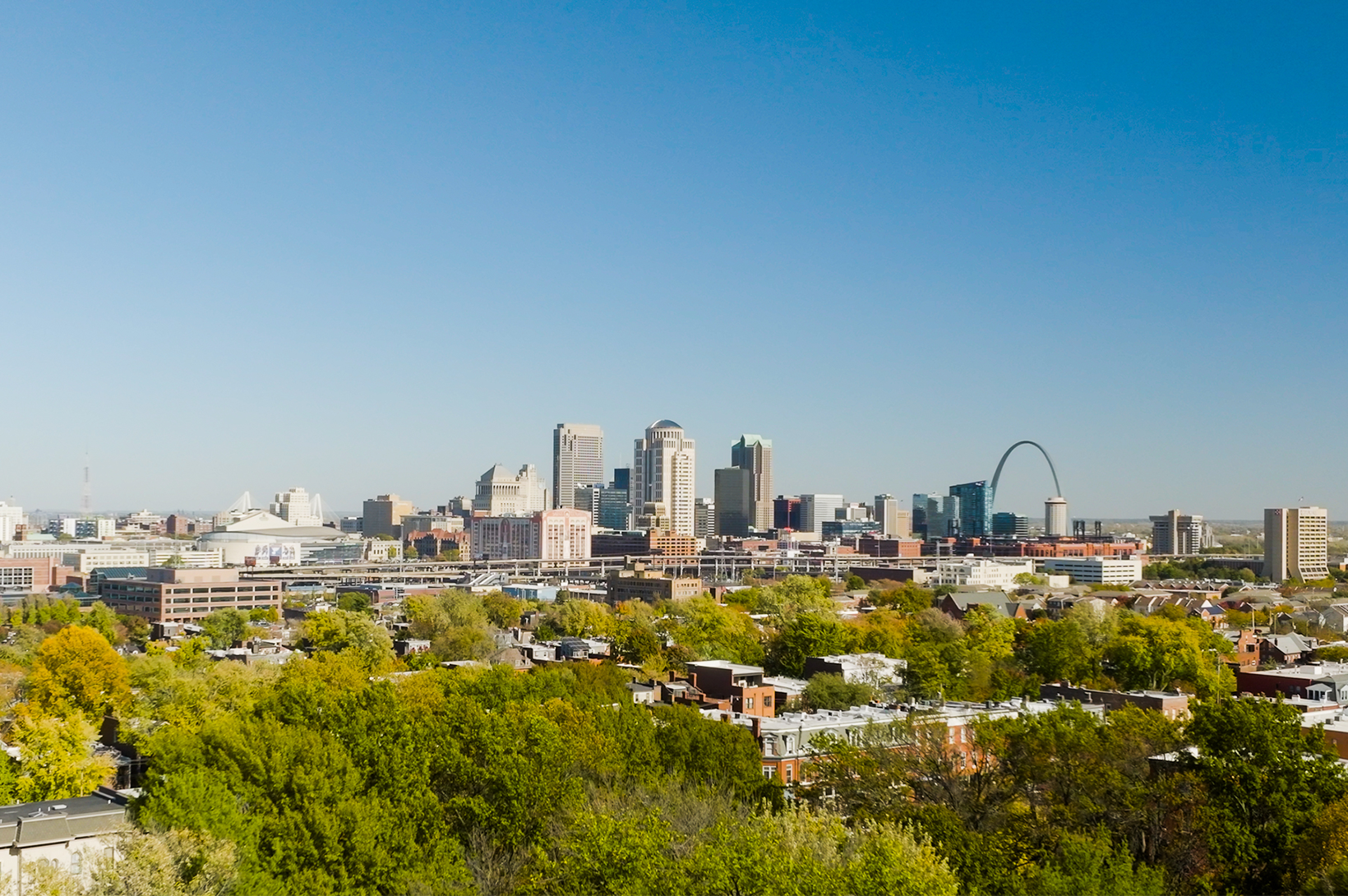 A panoramic photo of Downtown St. Louis taken from the south, featuring deep blue skies and greenery.