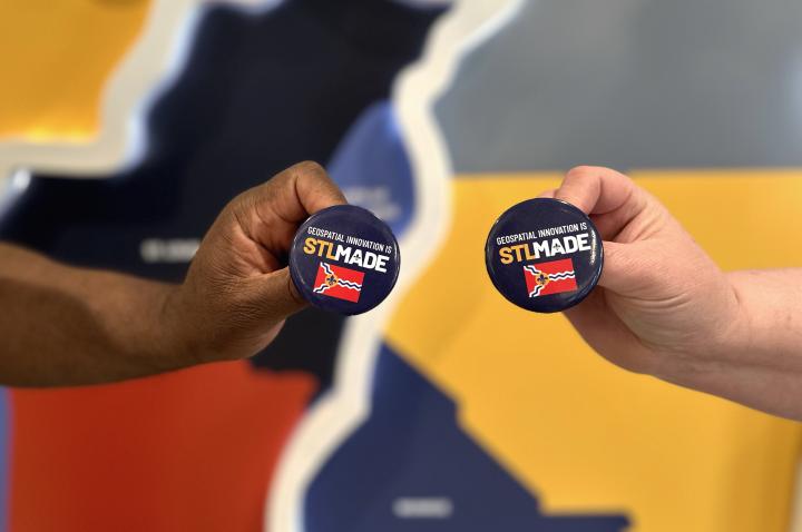 Two hands hold pins that say "Geospatial Innovation is STLMade" in front of a map of the St. Louis region.
