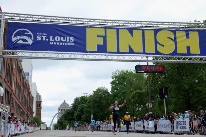 A photo of the finish line during the 2025 Greater St. Louis Marathon. A women runs victorious and the clock reads 3:54:56.