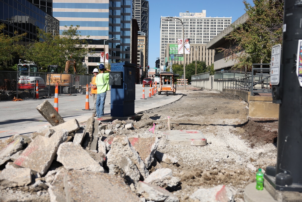 Urban street construction scene with broken concrete pile in foreground, workers in safety vests and orange cones near excavators