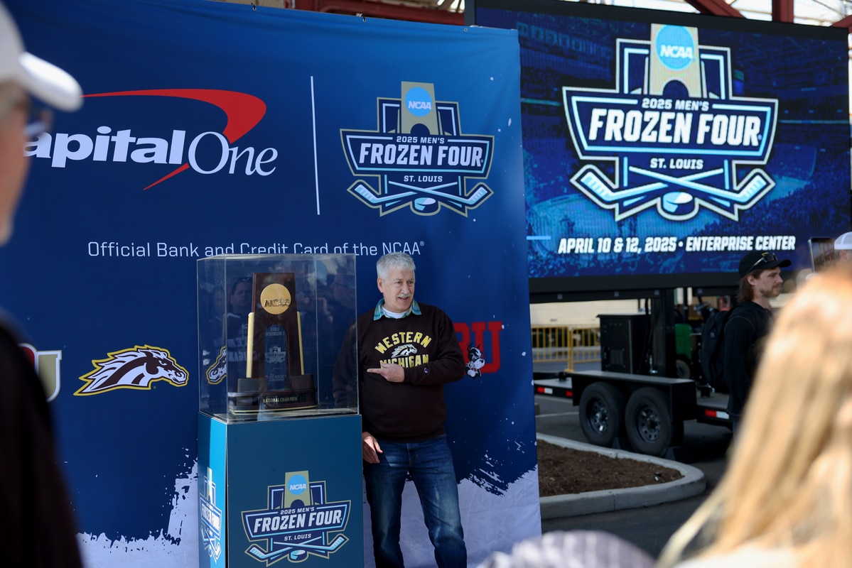 Man in a Western Michigan sweatshirt standing beside the NCAA Frozen Four trophy in a display case at a promotional booth with Frozen Four signage