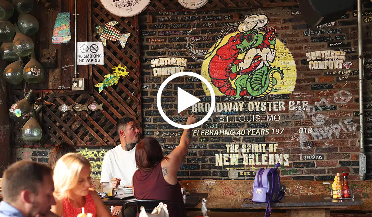 Casual seafood bar interior with diners at wooden tables in front of a graffiti-covered brick wall painted with a colorful red crawfish holding a green alligator and signage reading “Broadway Oyster Bar” and “The Spirit of New Orleans,” as a seated woman points toward the mural.