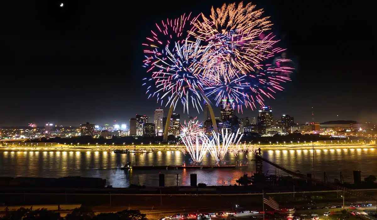 Colorful fireworks burst in pink, blue and gold over the illuminated Gateway Arch and downtown skyline at night, reflecting on the river with a crescent moon overhead.
