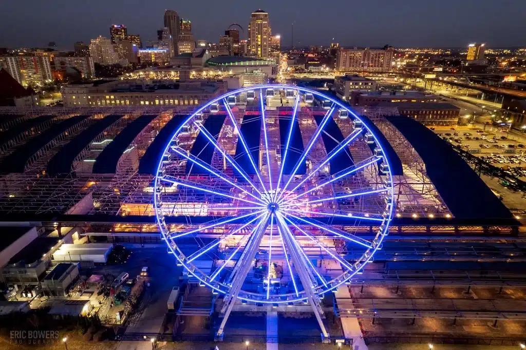 A large Ferris wheel lit with bright blue neon lights dominates the foreground above train tracks and station platforms, with a lit city skyline and buildings glowing against the dusk sky in the background.