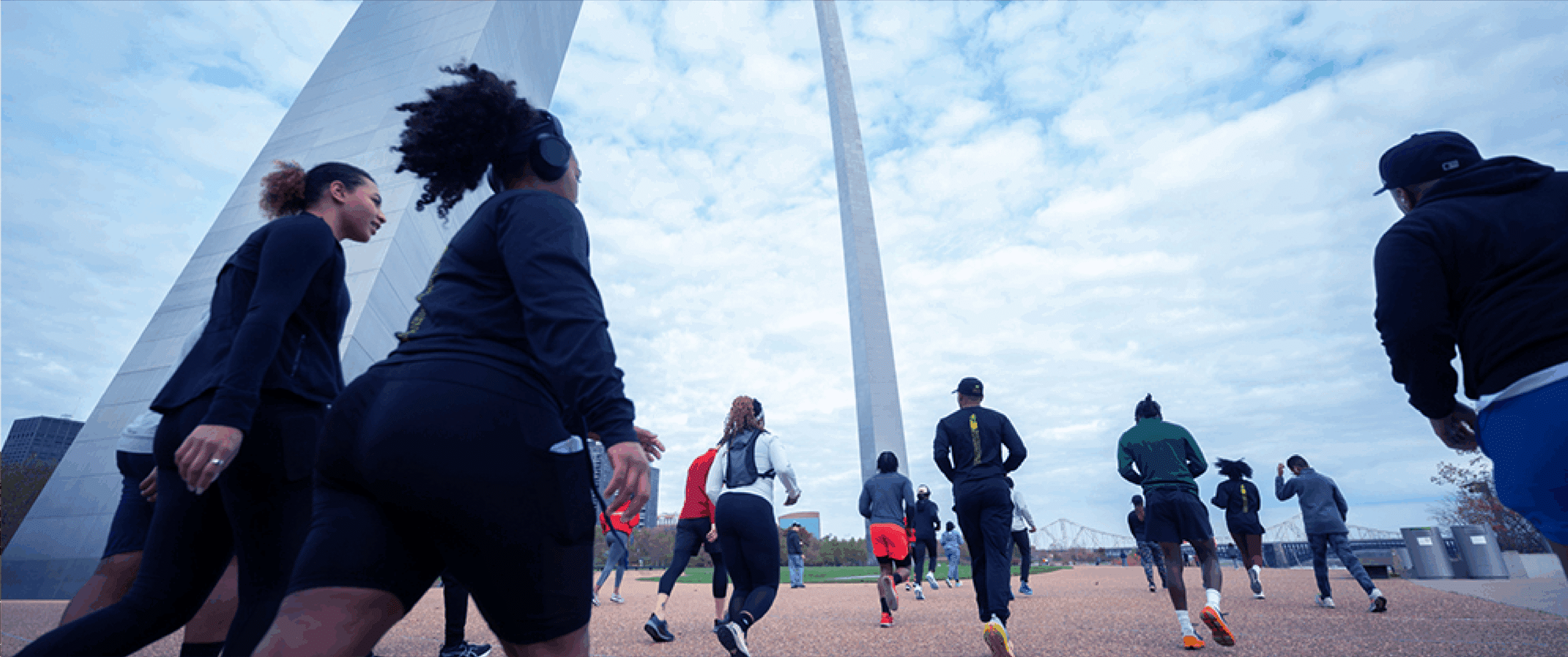 A group of runners in athletic clothing jog away from the camera toward a towering stainless-steel arch under a cloudy sky, captured from a low angle with the monument rising overhead.