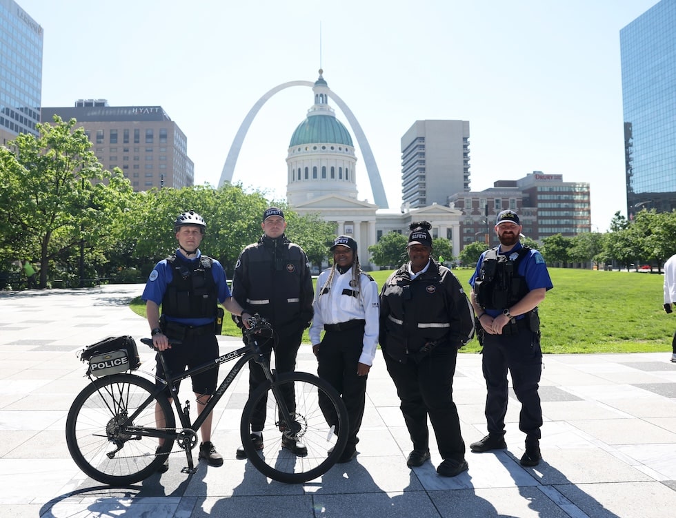 Five uniformed police and public-safety officers stand in a sunlit plaza with a marked police bicycle in front of them, the Old Courthouse dome and the Gateway Arch visible in the background.