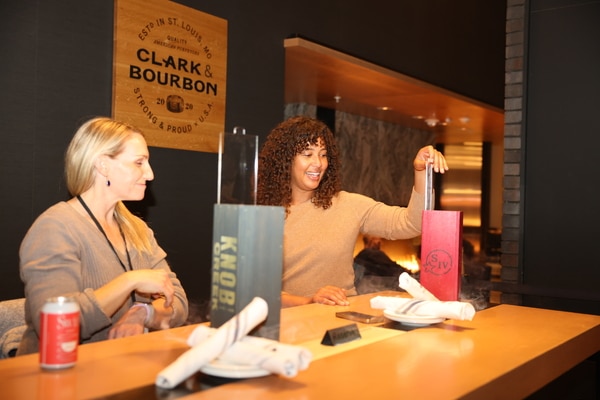 Rooftop swimming pool and sun deck with lounge chairs and a woven egg chair in the foreground, blue water bordered by a Greek-key tile band, and a city skyline under a bright, partly cloudy sky.
