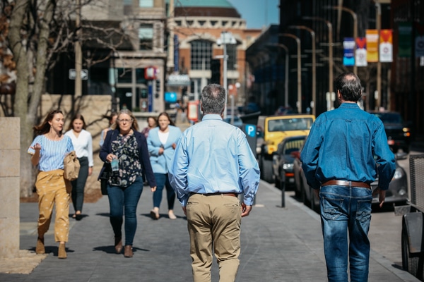 People on a city sidewalk; two men in blue shirts walk away from the camera while others approach, with parked cars nearby.