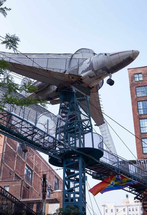 Retired narrow-body airplane perched atop a teal metal playground tower with enclosed wire-walkways between brick buildings.