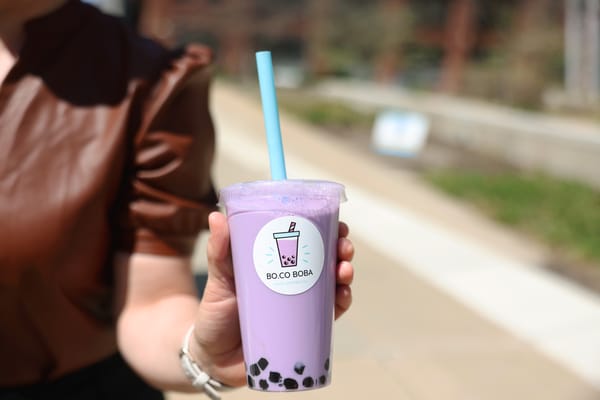 Neoclassical memorial building with tall rectangular columns, a carved dedication across the top, two flagpoles and stone soldier statues at the entrance, mirrored in a calm reflecting pool engraved with 'NAVY' and 'AIR FORCE', with a clear blue sky and city buildings in the background.