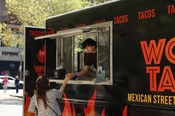 Red brick Victorian-style house with a rounded turret and arched porch behind a low stone wall, an American flag and flowering shrubs at the entrance under an overcast sky.
