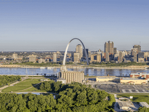Aerial view of the Gateway Arch towering over St. Louis's downtown skyline across the Mississippi River, with riverside warehouses and tree-lined parkland in the foreground.