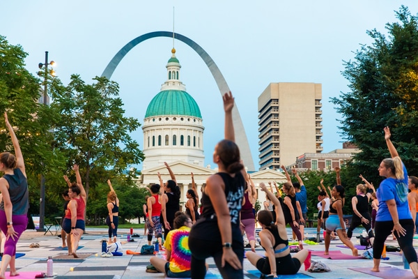 Rooftop bar at dusk with hanging string lights, people dining and lounging on curved sofas around a fire pit, a bartender at the bar, and a large arch and bridge on the city skyline.