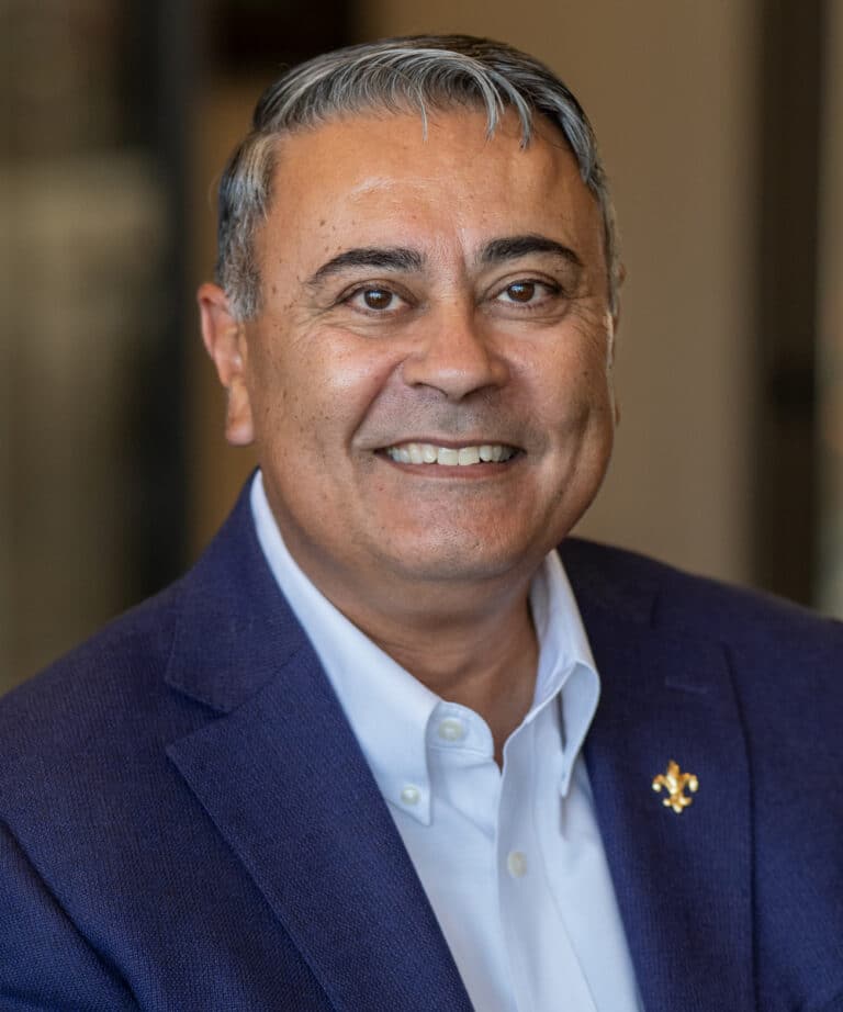 Smiling middle-aged man with salt-and-pepper hair wearing a blue blazer and white shirt with a gold fleur-de-lis lapel pin.