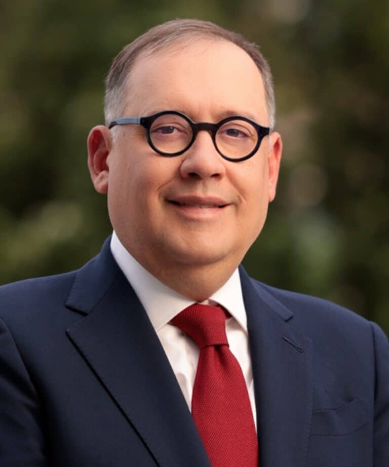 Middle-aged man in round black glasses wearing a navy suit, white shirt and red tie, smiling against a blurred green background.