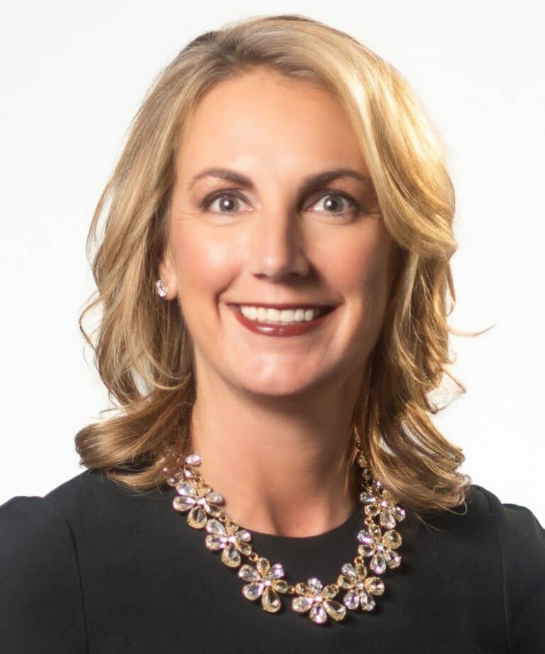 Smiling blonde woman in a black top wearing a sparkly floral necklace and stud earrings on a white background.