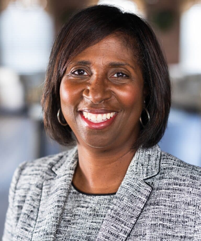 Smiling middle-aged woman with short dark hair wearing a gray patterned blazer and hoop earrings.