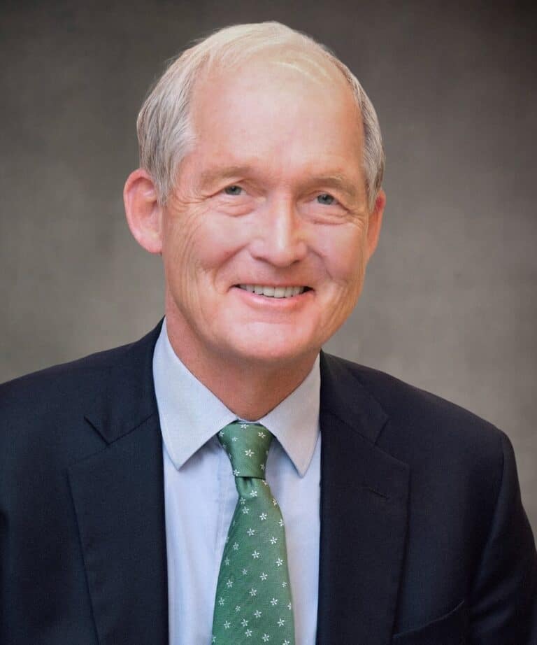 Smiling older man with gray hair wearing a dark suit, light blue shirt and green patterned tie against a gray background.