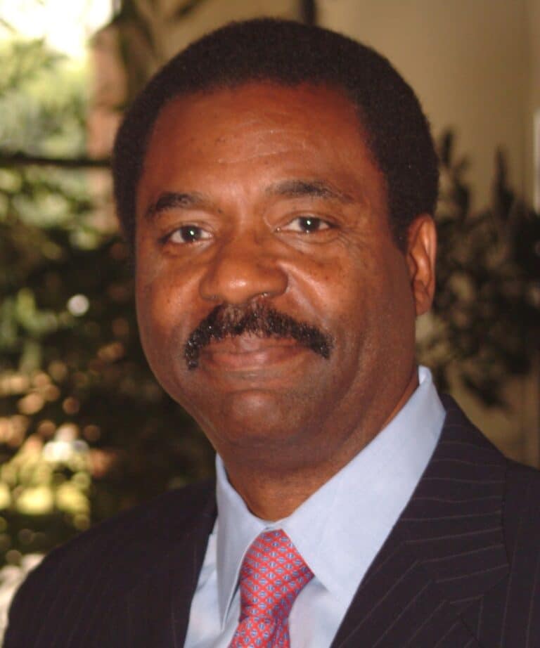 Middle-aged Black man with short hair and a mustache wearing a pinstripe suit, light blue shirt and red patterned tie, indoor plant background.