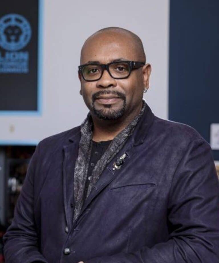 Bald man with glasses and trimmed goatee wearing a dark blazer, patterned scarf, and small hoop earring, standing indoors.