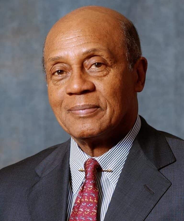 Bald older Black man in a dark suit and red patterned tie, facing camera with a slight smile against a gray backdrop.