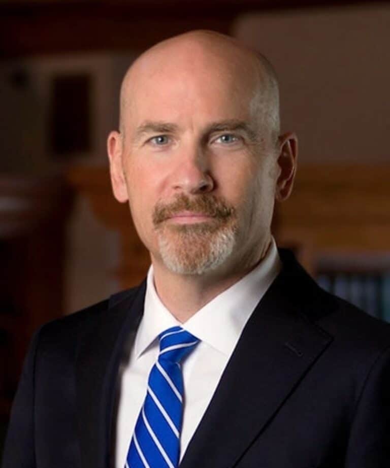 Bald man with trimmed goatee wearing a dark suit, white shirt and blue striped tie, standing against a blurred office background.