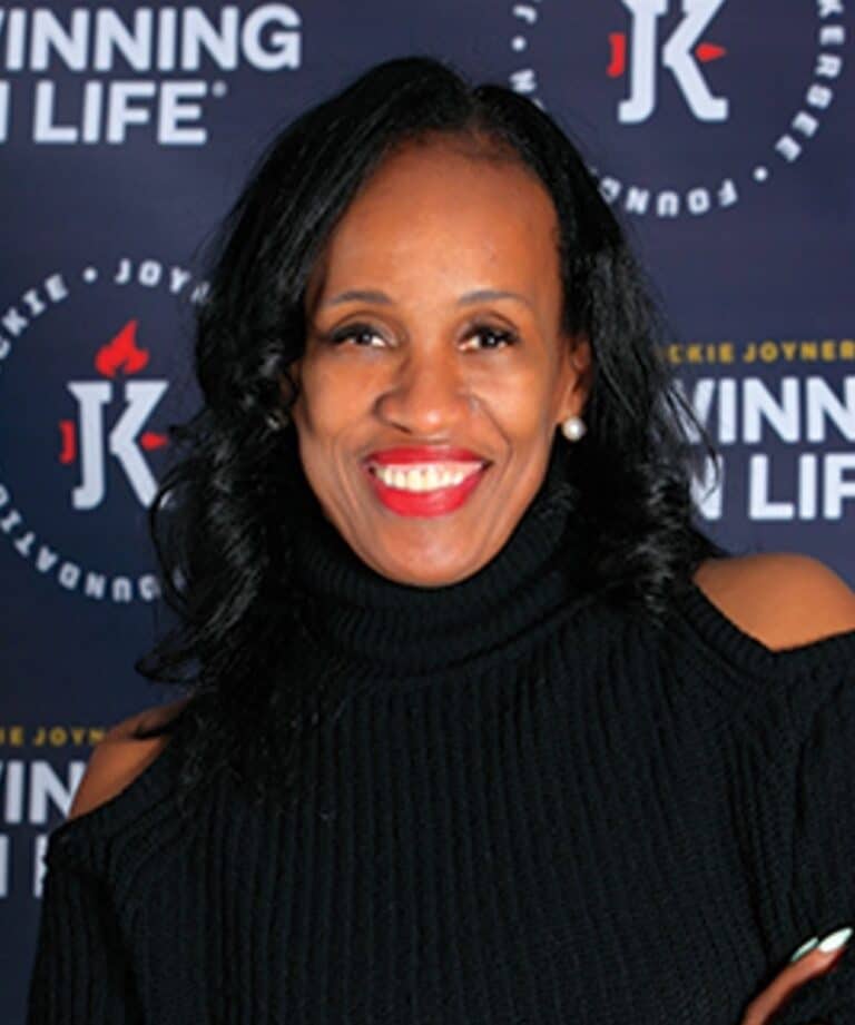 Smiling woman with wavy dark hair, red lipstick and pearl earring wearing a black cut-out turtleneck against a logo backdrop.