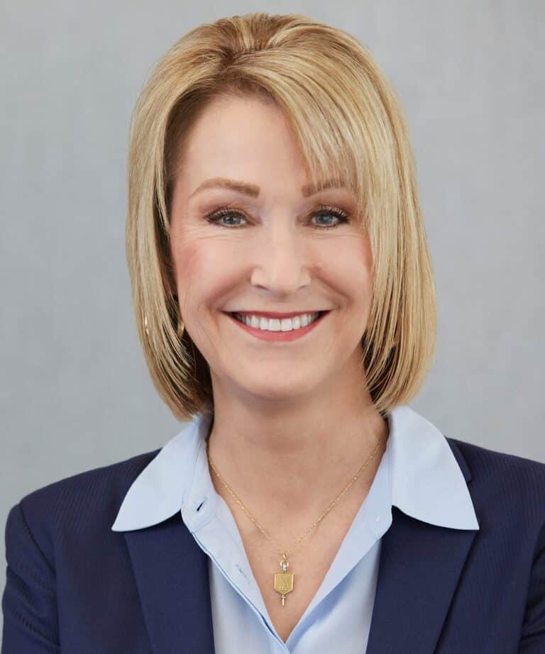 Smiling woman with short blonde bob wearing a navy blazer, light-blue shirt and gold pendant necklace.