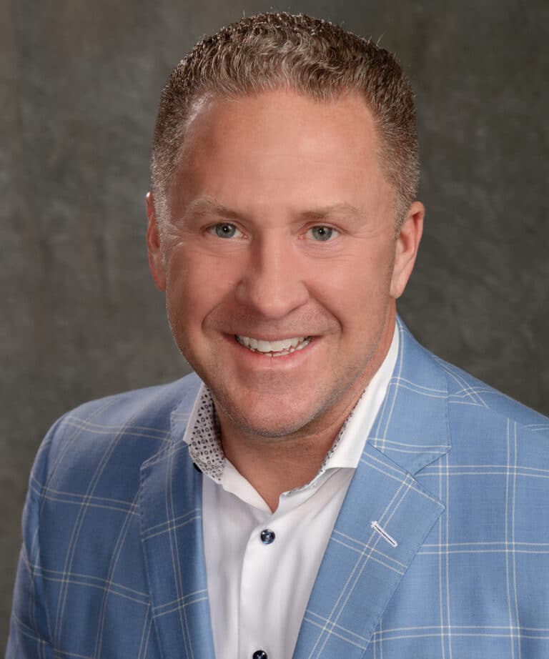 Smiling middle-aged man with short curly brown hair wearing a blue plaid blazer and white shirt against a gray backdrop.