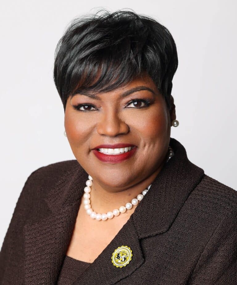 Smiling woman with short black hair, red lipstick, pearl necklace and earrings, wearing a dark blazer with yellow lapel pin.