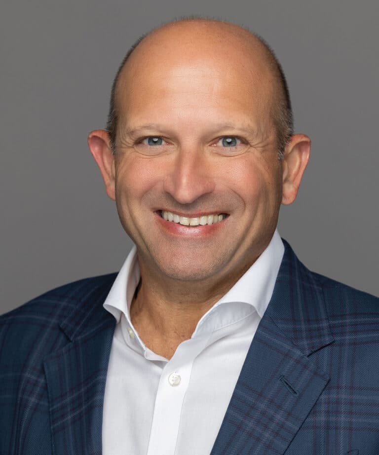 Bald man in a blue plaid suit jacket and white shirt smiling against a gray background.