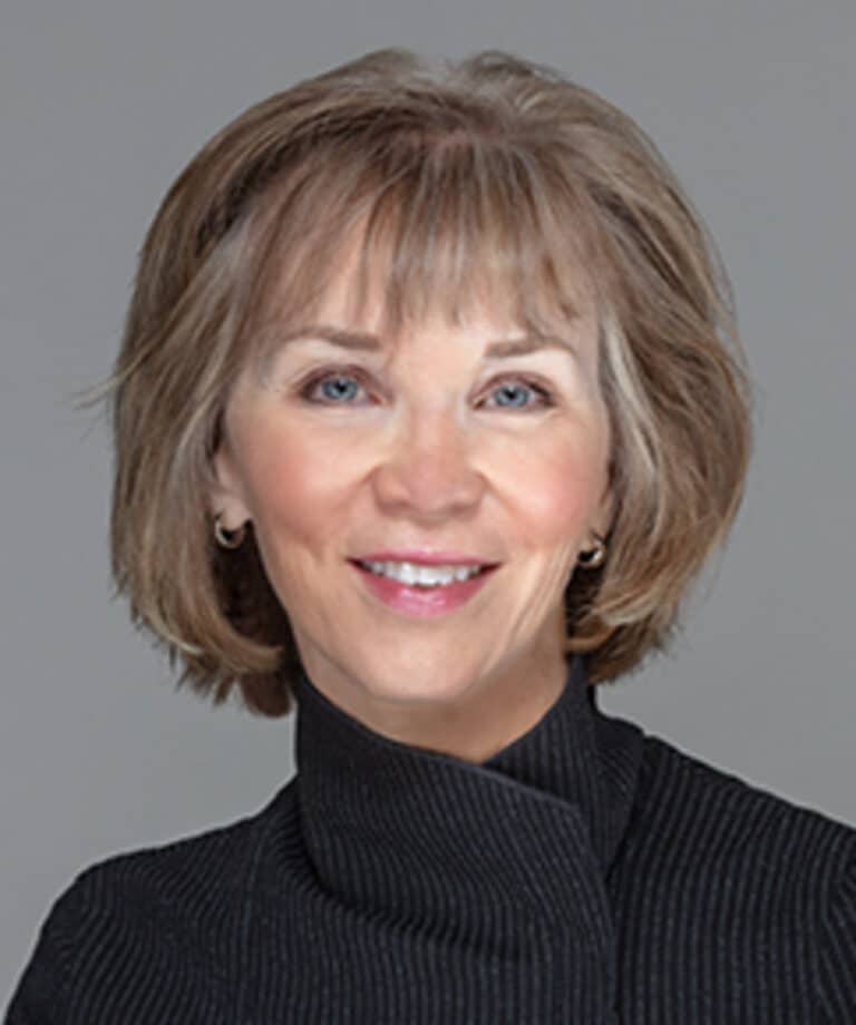 Smiling middle-aged woman with short light brown hair and bangs, blue eyes, small hoop earrings, wearing a dark high-collared jacket.