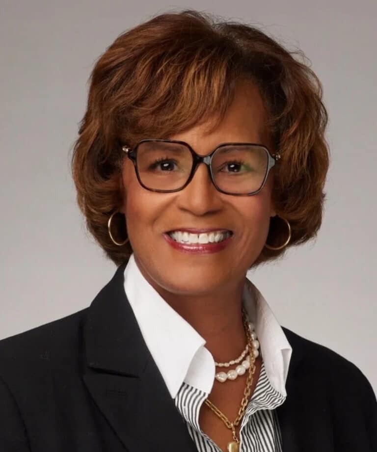 Smiling woman with short brown hair and glasses wearing hoop earrings, layered pearl and gold necklaces and a black blazer.