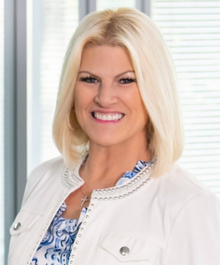 Smiling blonde woman with short hair wearing a white studded jacket, patterned blouse, and cross necklace in an office.