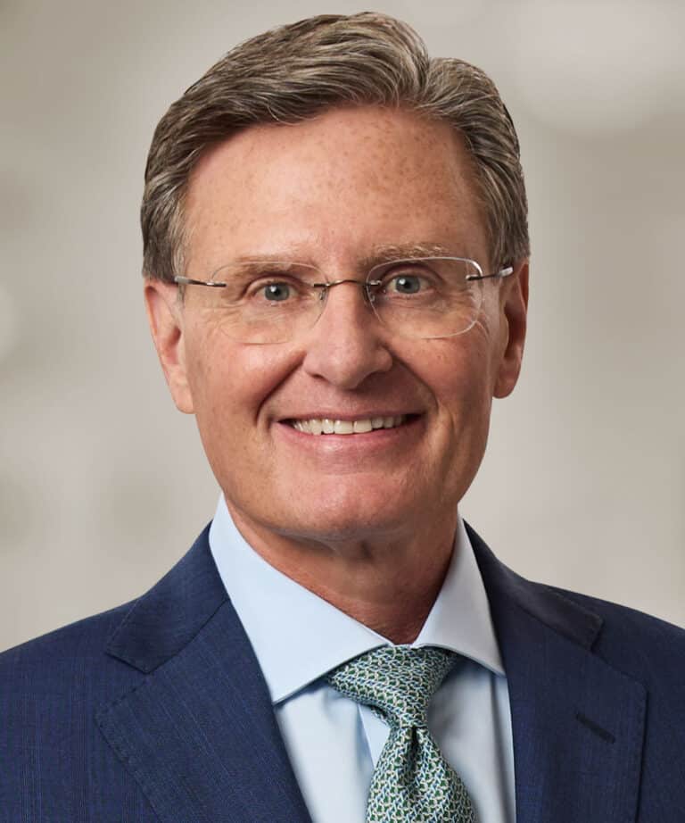 Smiling older man with gray hair and thin metal-frame glasses wearing a blue suit, light blue shirt, and patterned green tie.
