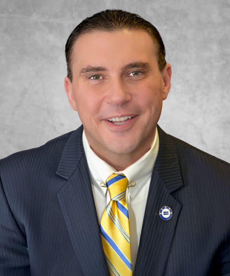 Smiling man in a dark pinstripe suit, white shirt, yellow-and-blue striped tie, round blue lapel pin, gray textured backdrop