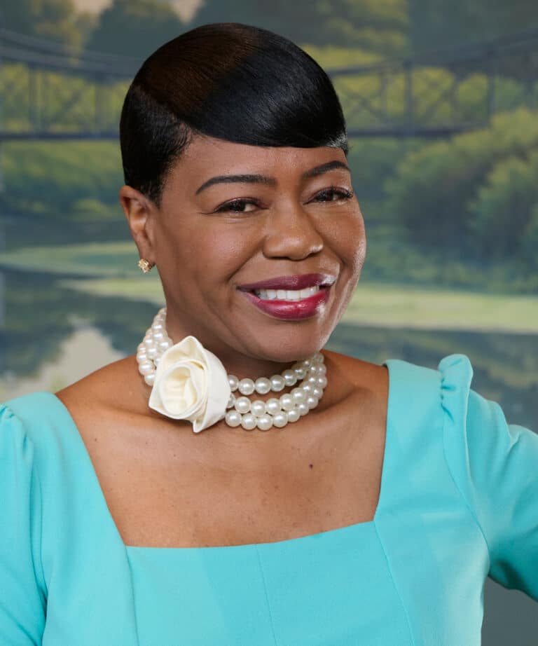 Smiling woman with short sleek hair wearing a turquoise square-neck dress and a triple-strand pearl choker with a white rose.