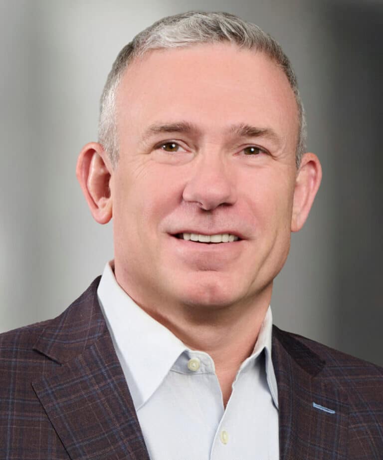Middle-aged man with short gray hair, smiling, wearing a dark checked suit jacket and white collared shirt