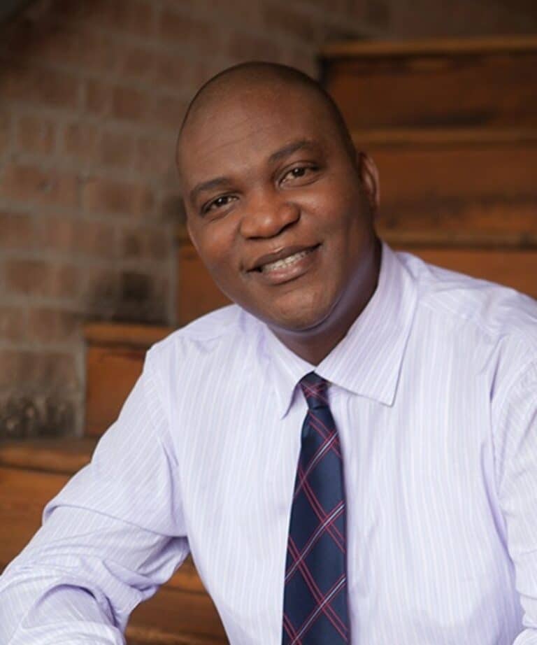 Smiling bald man in a light striped dress shirt and patterned tie seated on wooden stairs against a brick wall.