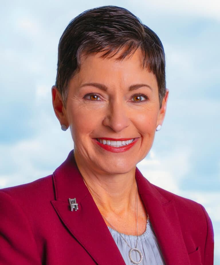 Smiling middle-aged woman with short dark hair, red lipstick, stud earrings, maroon blazer and circular pendant necklace.