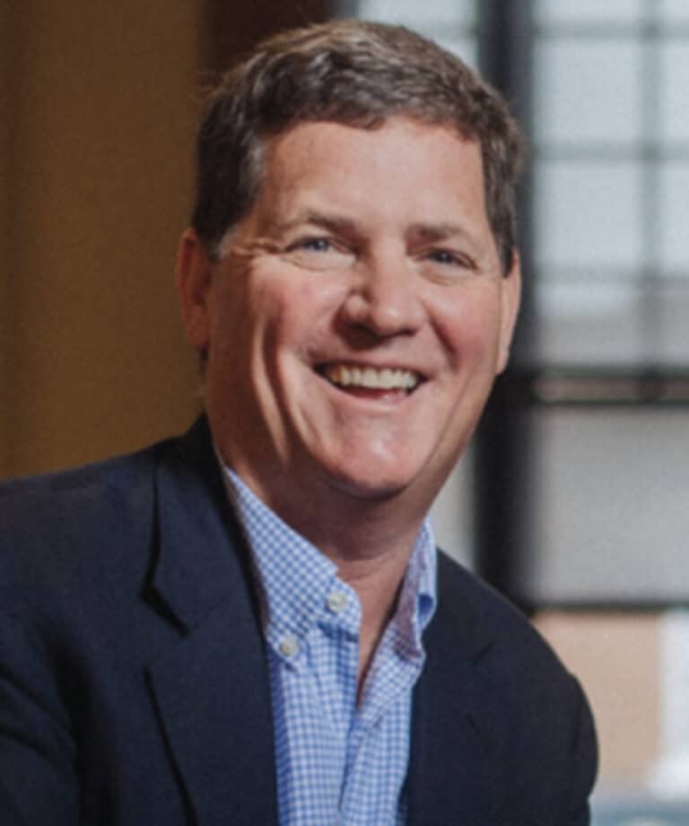 Smiling middle-aged man with short brown hair wearing a navy blazer and blue checkered shirt in an indoor setting