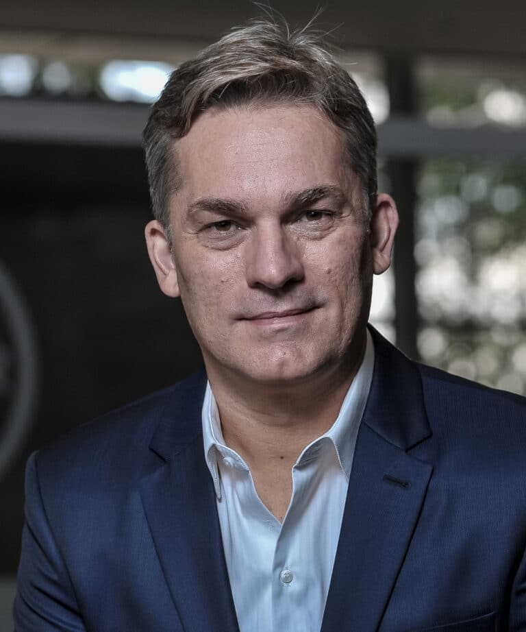 Middle-aged man in a navy suit and white shirt with a neutral expression, close-up headshot and blurred background.