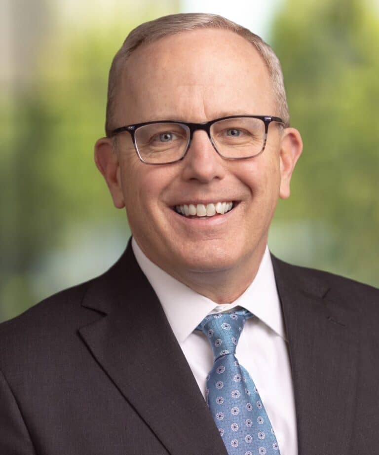 Smiling man with glasses and short light hair in a dark suit and blue patterned tie against a blurred green background.