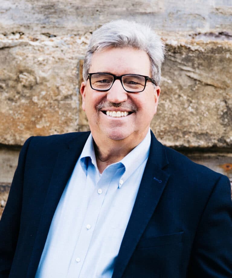 Smiling middle-aged man with gray hair, mustache and glasses wearing a navy blazer and light-blue shirt against a stone wall.
