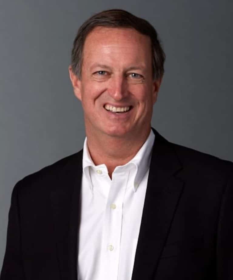 Smiling middle-aged man with short brown hair wearing a dark blazer and white shirt against a plain gray background.