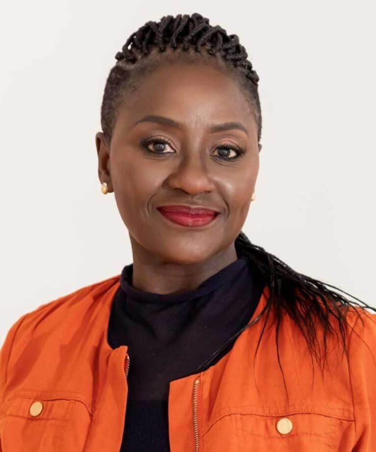 Smiling woman with braided hair pulled back, wearing an orange jacket over a black top, gold stud earrings and red lipstick.