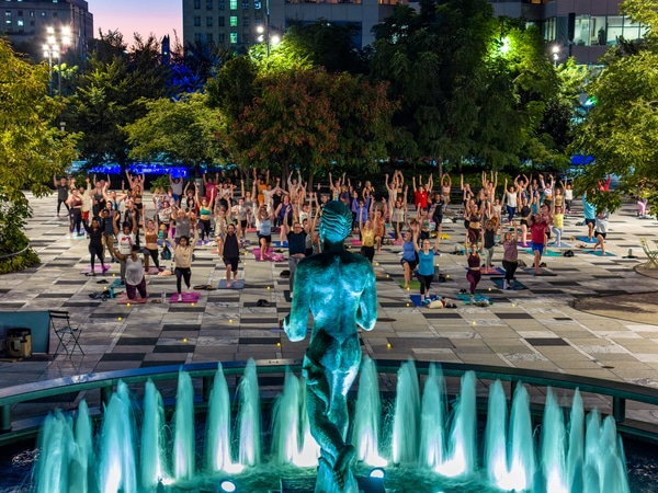 Neoclassical memorial building with tall rectangular columns, a carved dedication across the top, two flagpoles and stone soldier statues at the entrance, mirrored in a calm reflecting pool engraved with 'NAVY' and 'AIR FORCE', with a clear blue sky and city buildings in the background.